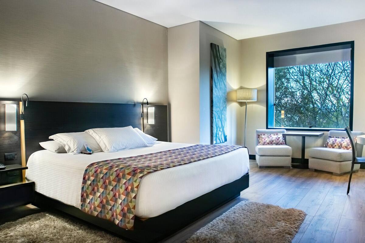 A modern bedroom with a large bed, bedside lamps, a desk, and a view of trees through a window.