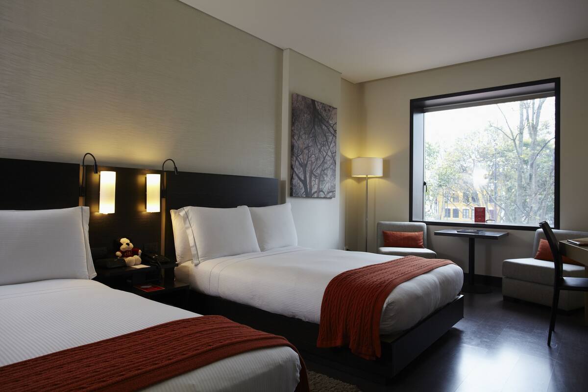 A hotel room with two beds, a desk, a chair, a window with a view, and a wall-mounted artwork.