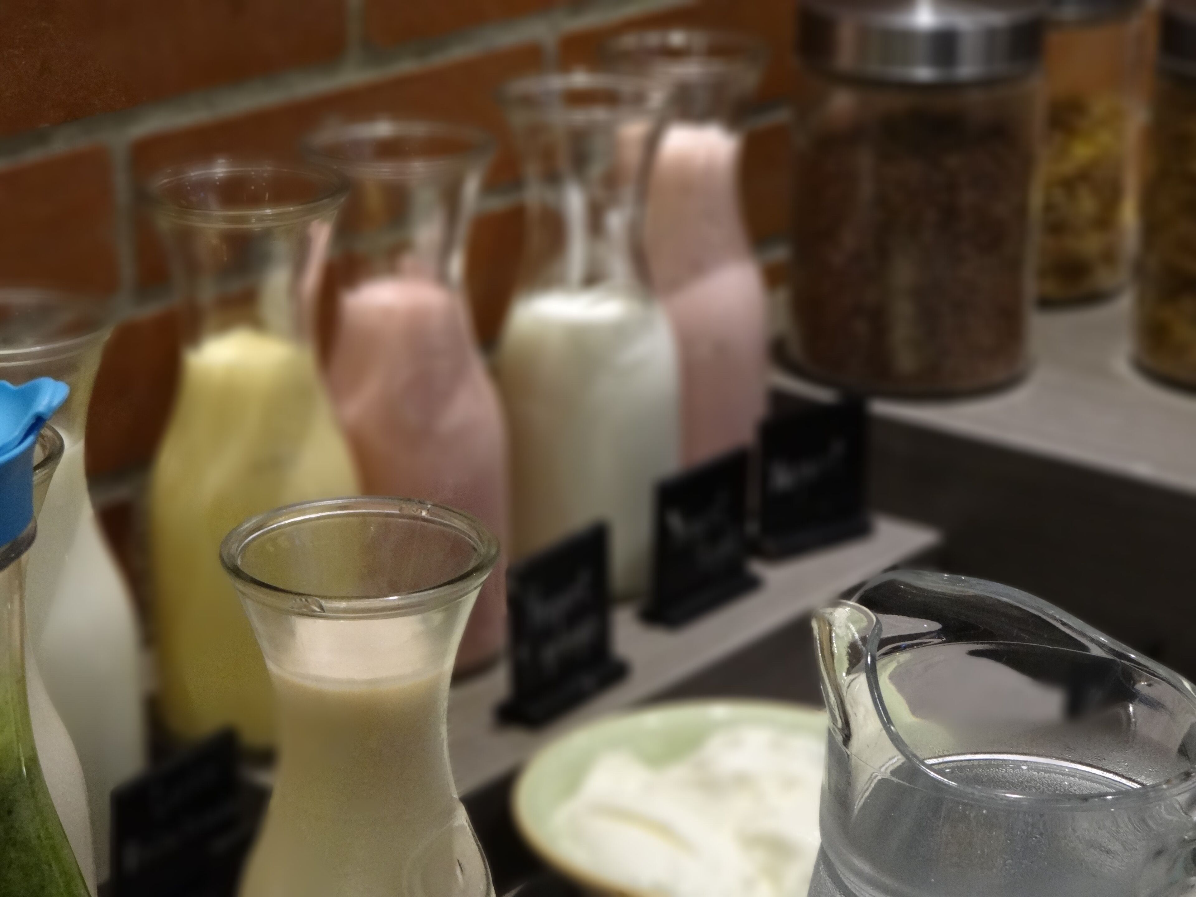 A selection of milkshakes in various flavors displayed in clear glass bottles with labels, set against a brick wall background.