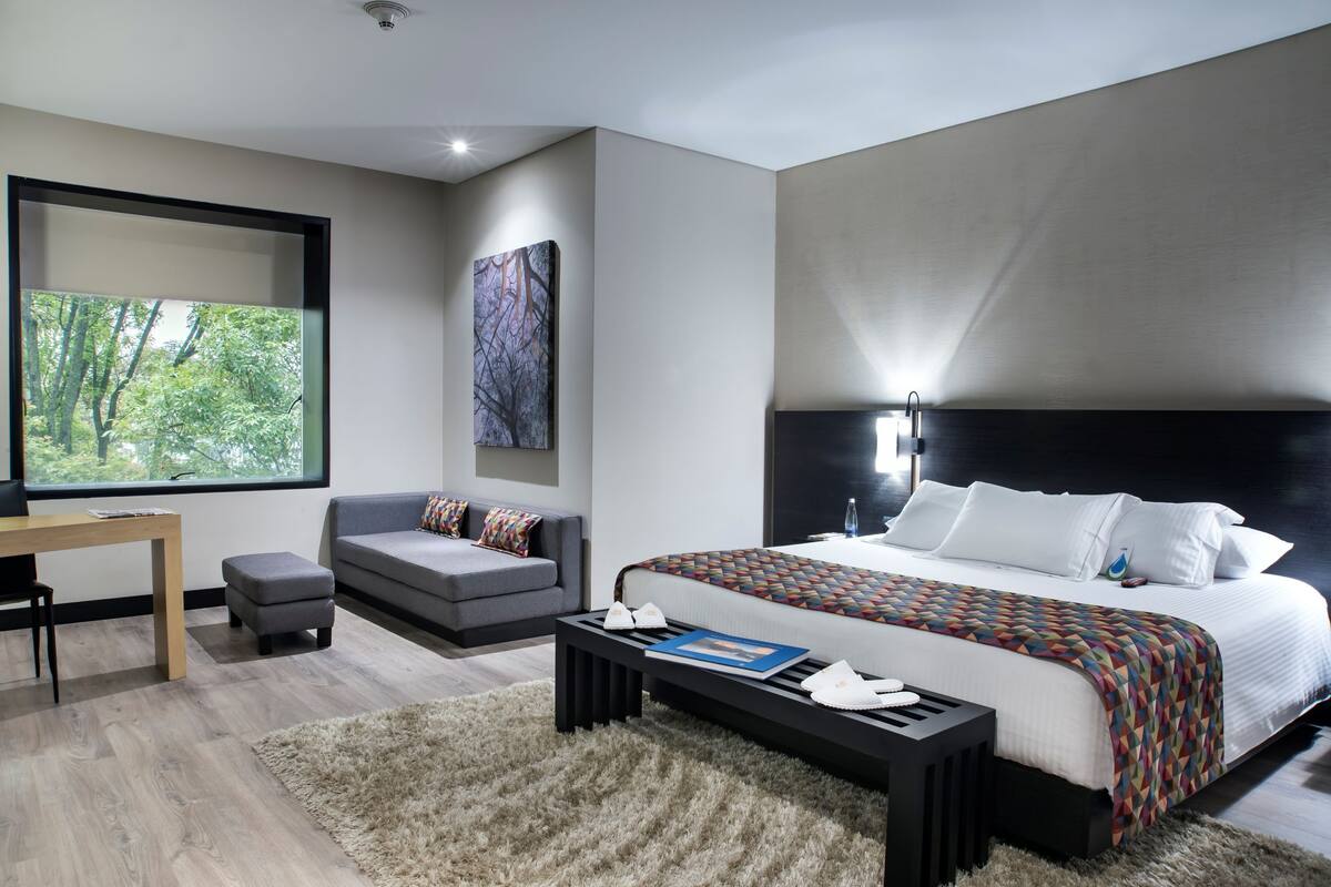 A modern bedroom with a large bed, a desk, a sofa, and a view of greenery through a window.