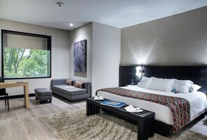 A modern bedroom with a large bed, a desk, a sofa, and a view of greenery through a window.