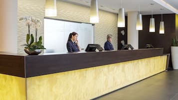 A reception desk with a modern design, featuring a dark wood finish and a yellow patterned section. There are three receptionists seated behind the desk, and a large potted plant is visible on the left.