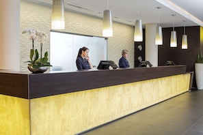 A reception desk with a modern design, featuring a dark wood finish and a yellow patterned section. There are three receptionists seated behind the desk, and a large potted plant is visible on the left.