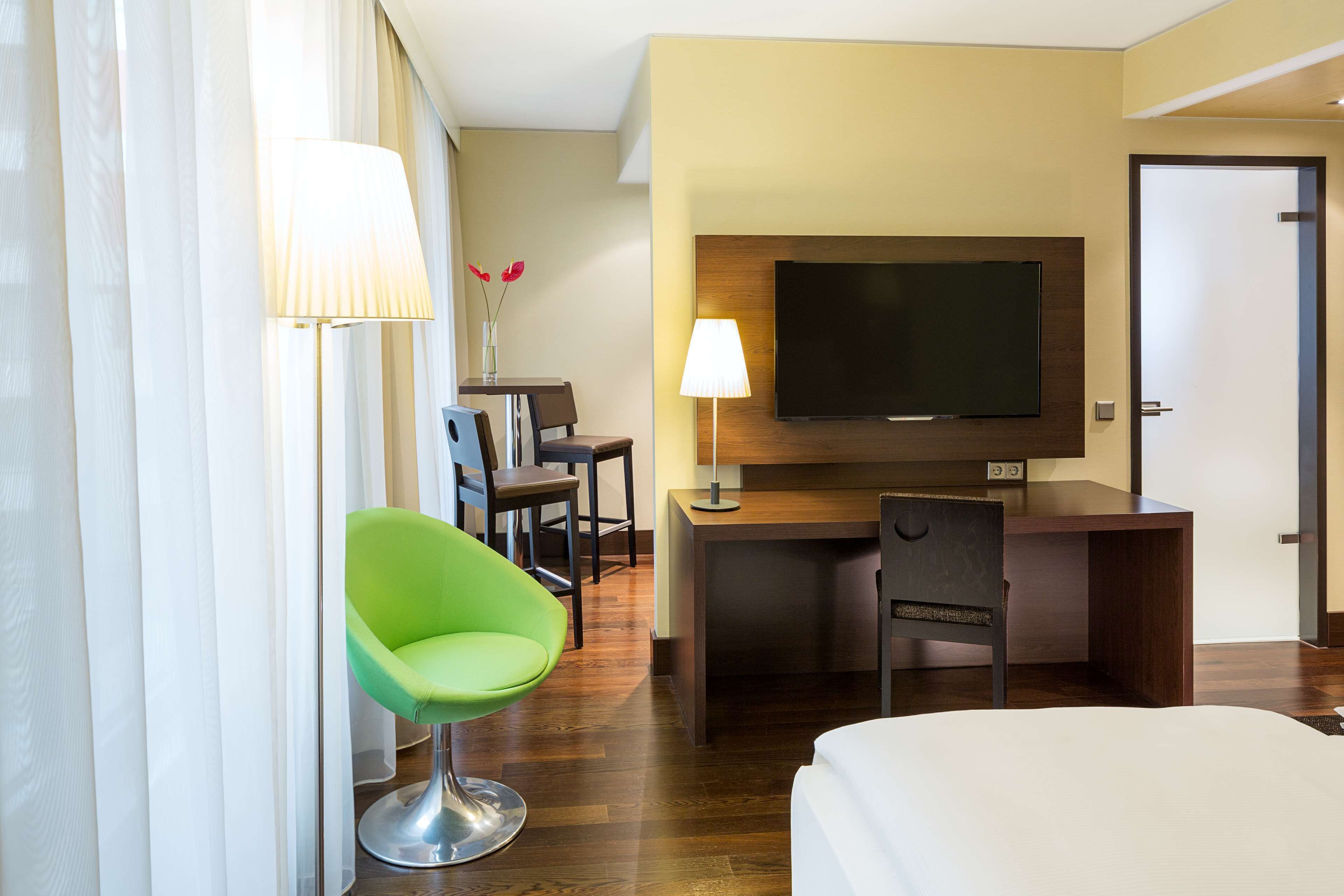 A hotel room with a bed, a desk with a TV, a green chair, and a lamp.