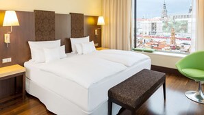 A hotel room with a large bed, two bedside lamps, a bench, a green chair, and a view of the city through a large window. - NH Collection Dresden Altmarkt (Dresden)