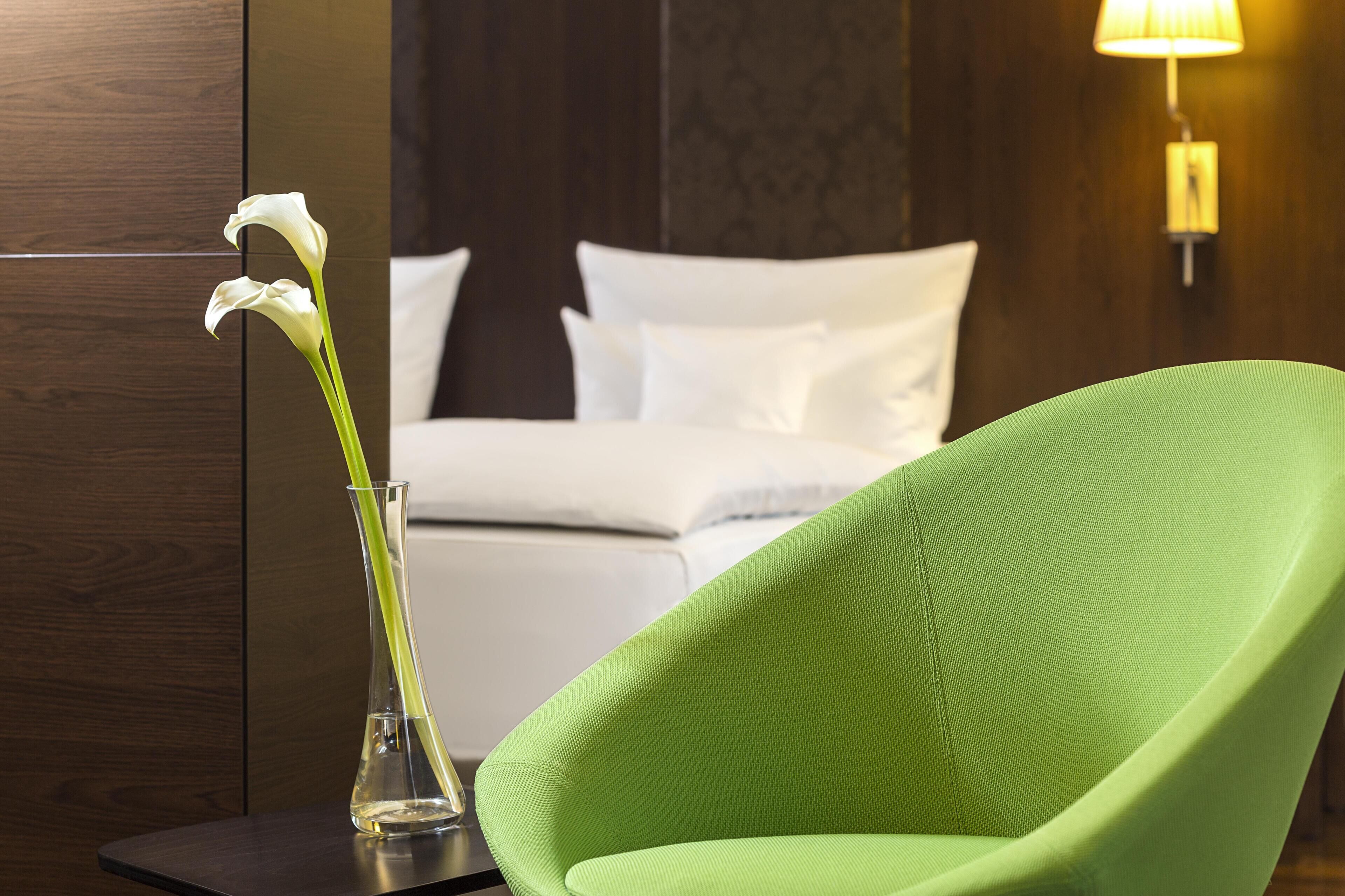 Una habitación con una silla verde, una cama con ropa de cama blanca y un jarrón con flores.