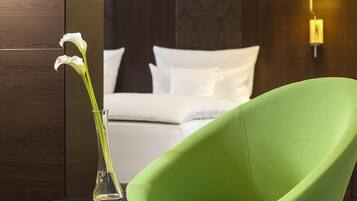 Una habitación con una silla verde, una cama con ropa de cama blanca y un jarrón con flores.