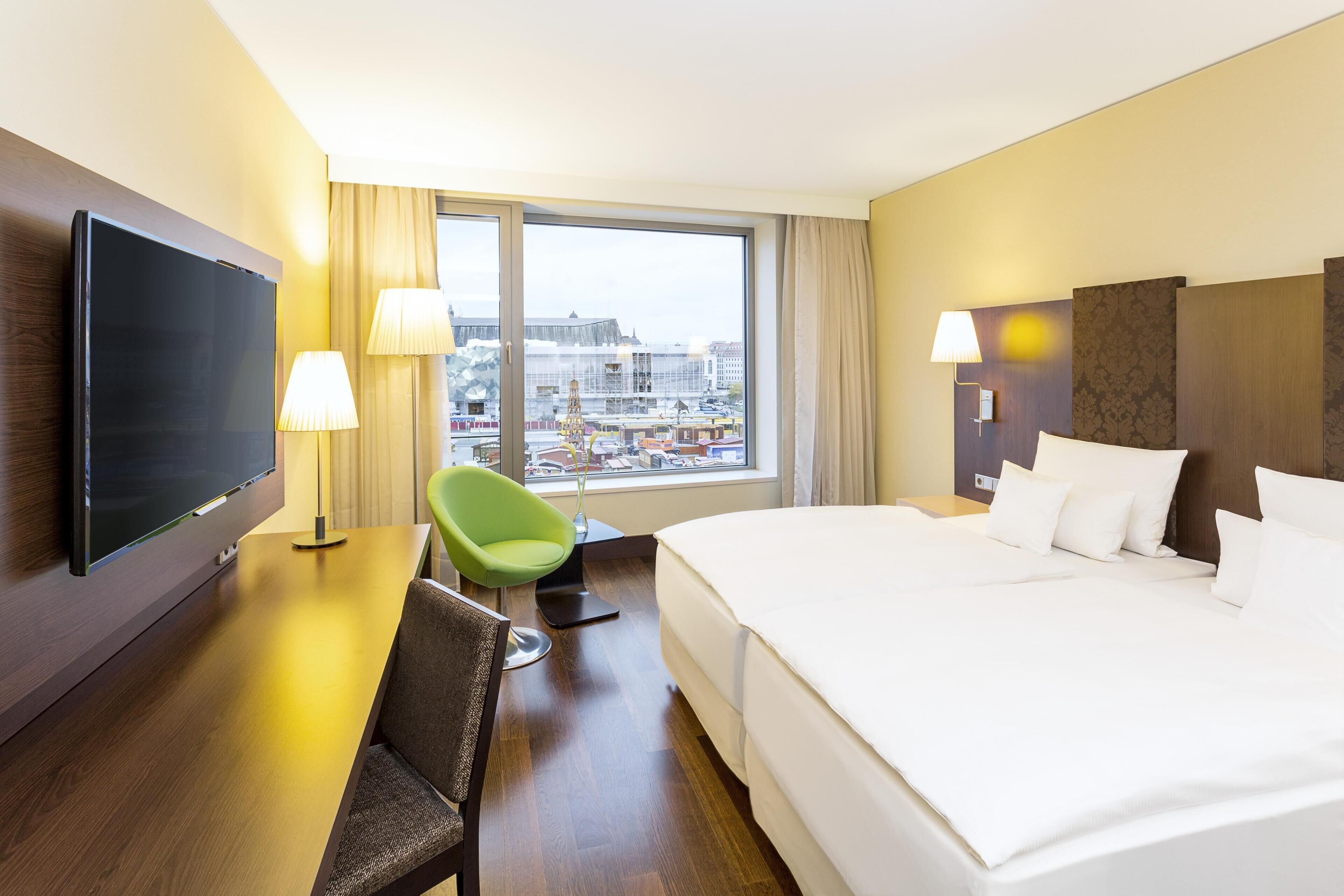 A hotel room with a large bed, a flat-screen TV, a desk with a chair, and a view of the city through large windows.