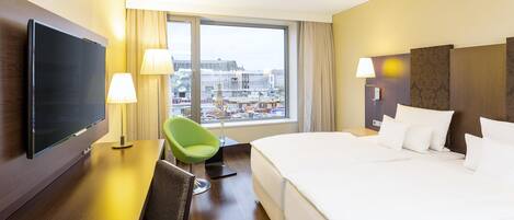 Habitación de hotel con una cama grande, televisión de pantalla plana, escritorio con silla y vistas a la ciudad a través de amplios ventanales.