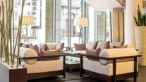 A modern lounge area with two sofas, a glass coffee table, and a large window offering a view of a historic building. - NH Collection Dresden Altmarkt (Dresden)