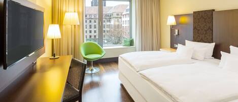 Une chambre d’hôtel avec un grand lit, un bureau doté d’un téléviseur à écran plat, un fauteuil vert et une fenêtre donnant sur des immeubles.