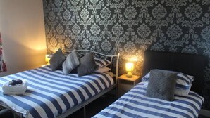A hotel room with two beds, each with striped bedding and pillows. There are bedside lamps and a small table with a towel and a bag on it. The walls have a patterned wallpaper. - The Vale Hotel (Hull)
