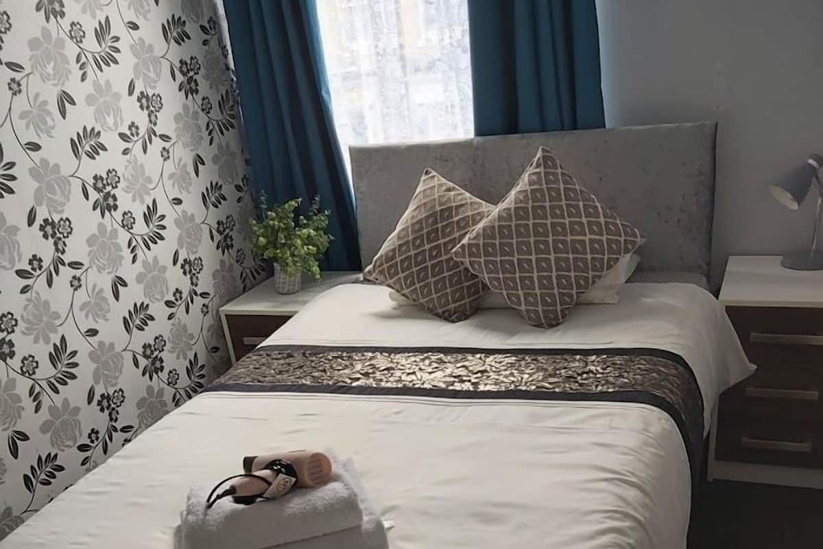A neatly made bed with a white duvet, a pair of slippers, and two patterned pillows.