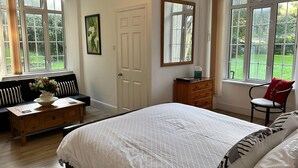 A bedroom with a large bed, a wooden dresser, a bench with cushions, a window with a view of greenery, and a door. - Exmoor Manor Guest House (Lynton)