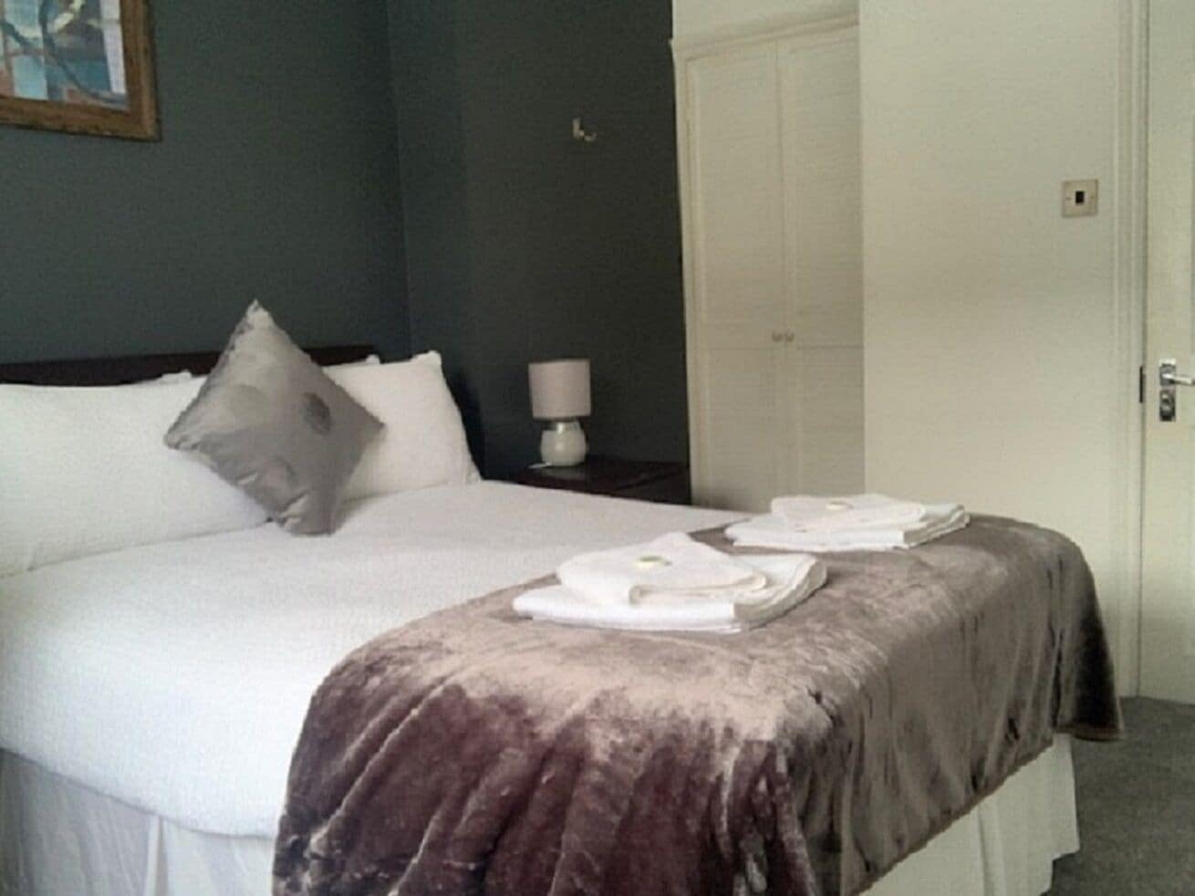 Una cama bien hecha con edredón blanco y manta marrón, una almohada y una toalla doblada sobre la cama.