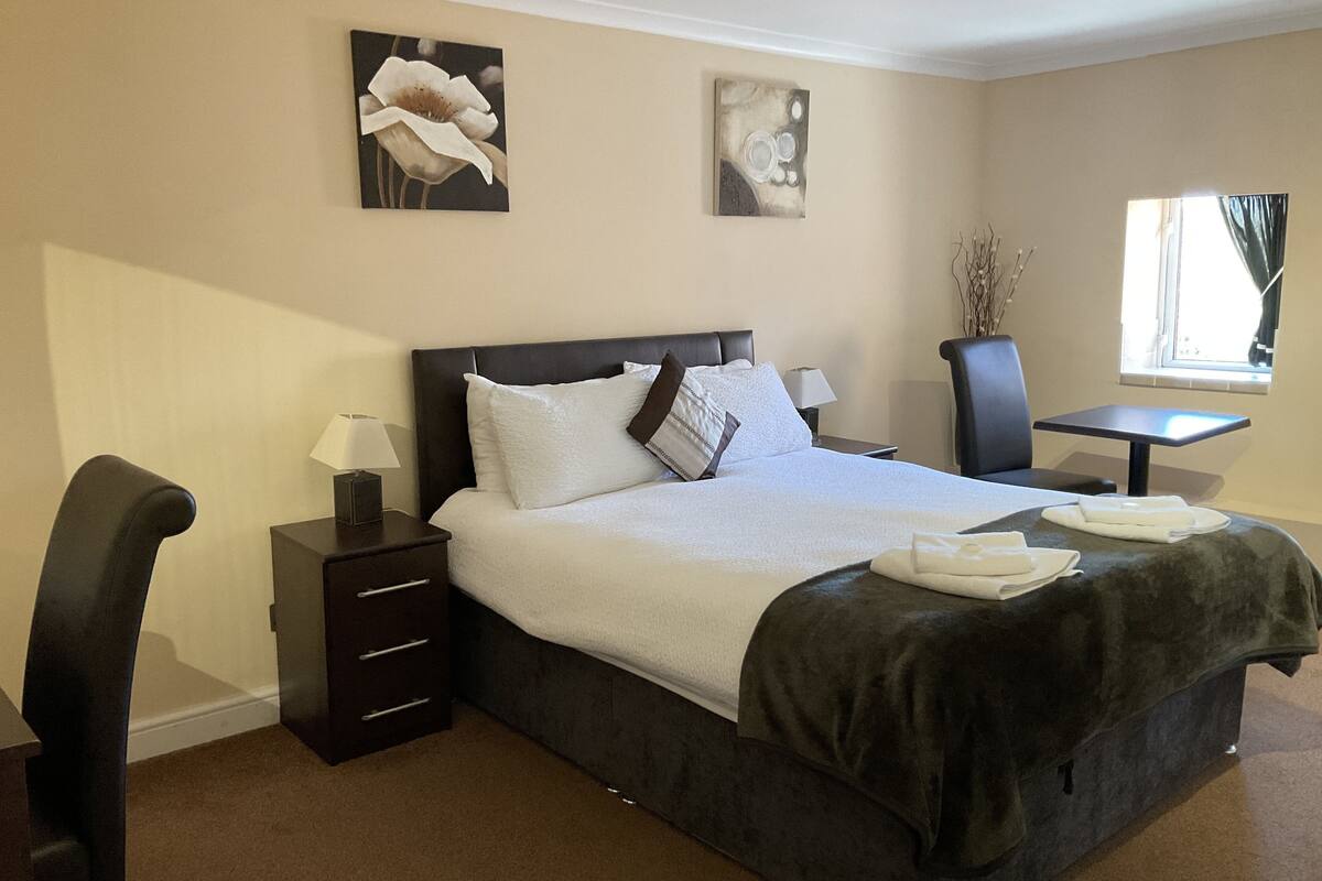A hotel room with a large bed, a bedside table with a lamp, a chair, a small table, and two wall-mounted paintings.