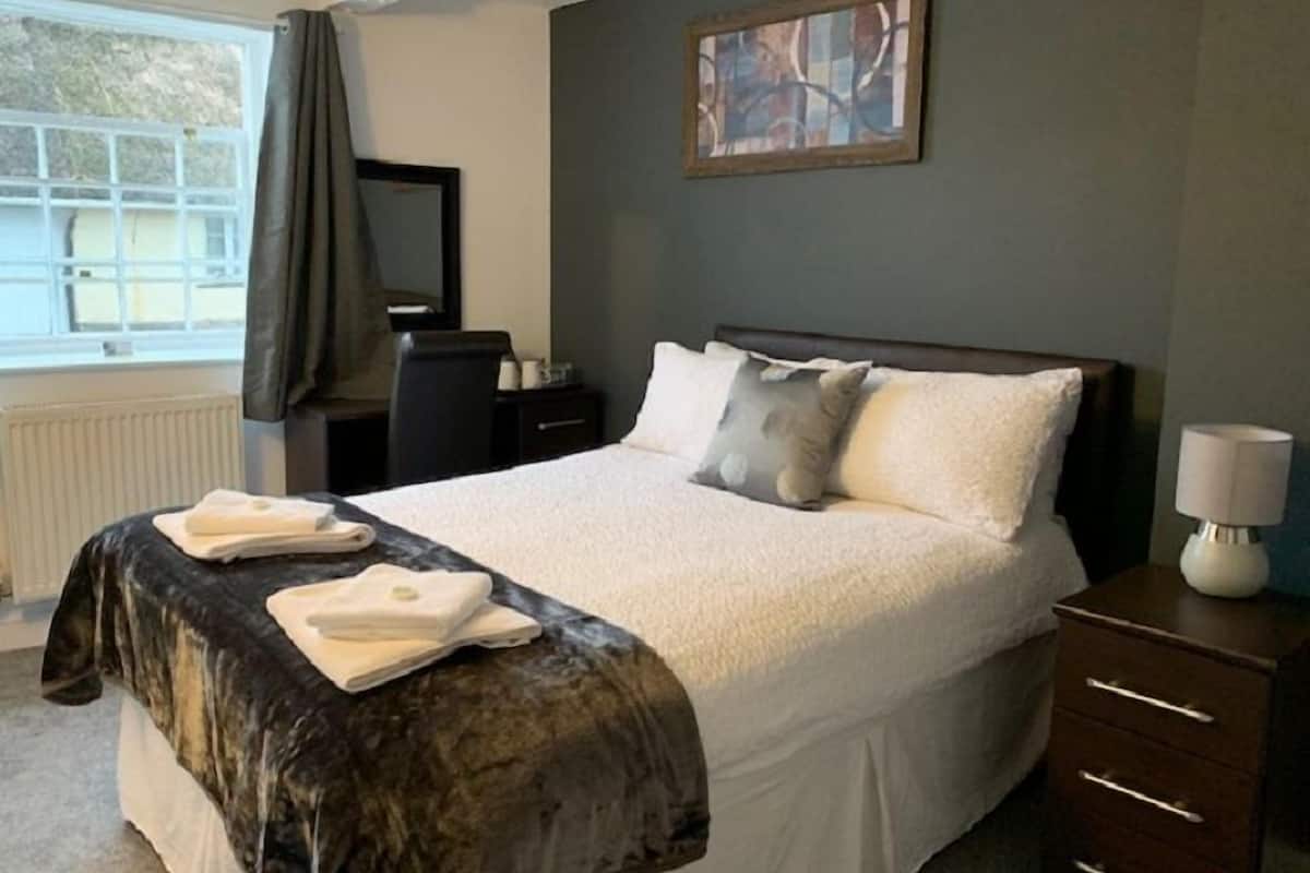 A neatly made bed with a white comforter and pillows, a bedside table with a lamp, a window with curtains, and a painting on the wall.