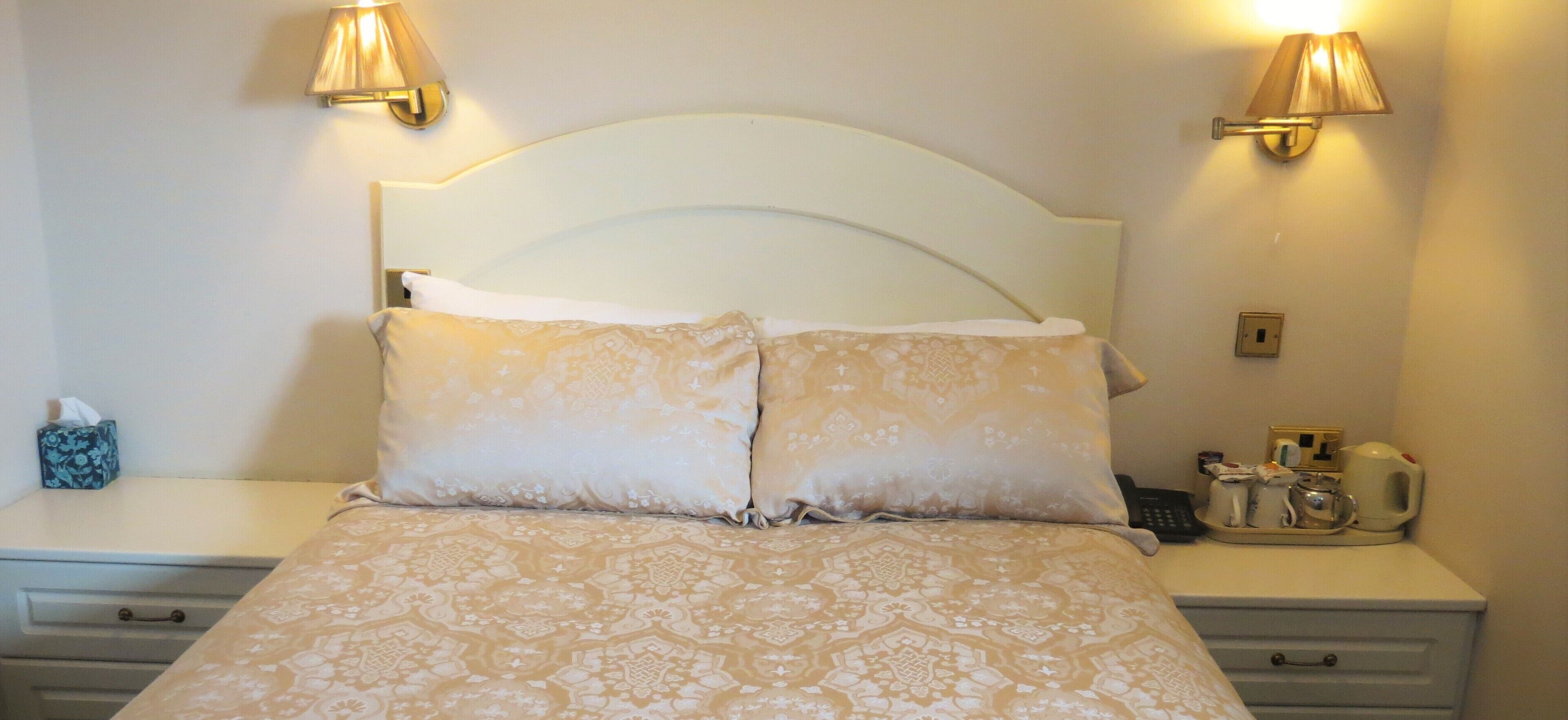 A neatly made bed with patterned bedding, two bedside lamps, a bedside table with a tissue box, a cup, and a kettle.