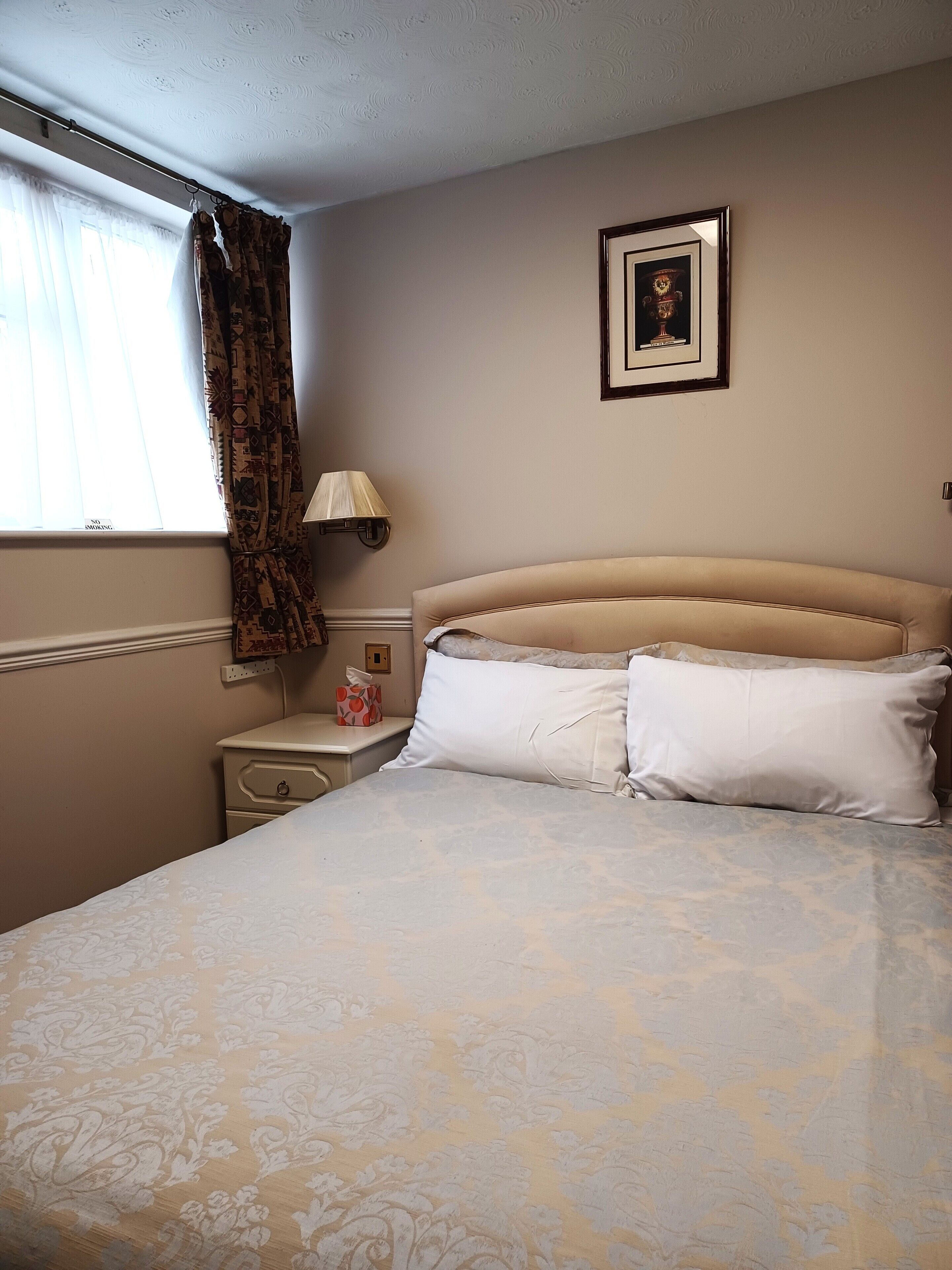 A bedroom with a bed, a nightstand, a lamp, a window with curtains, and a framed picture on the wall.