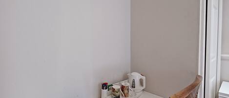 A corner of a room with a white desk, a wicker chair, a framed picture, and a tray with a mug and a kettle.