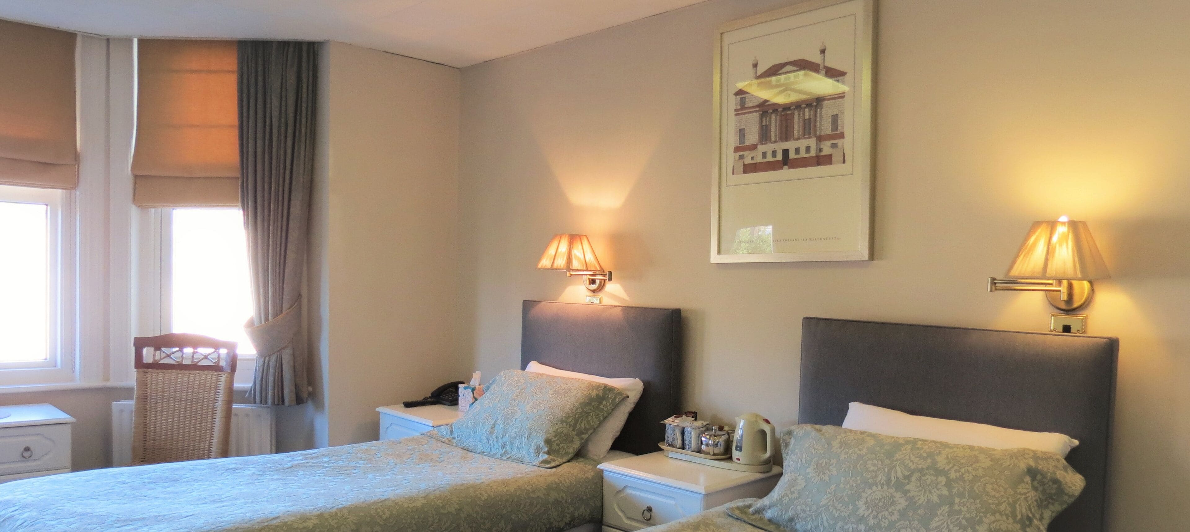 A hotel room with two beds, nightstands, lamps, a radiator, a wicker chair, and a framed picture on the wall.