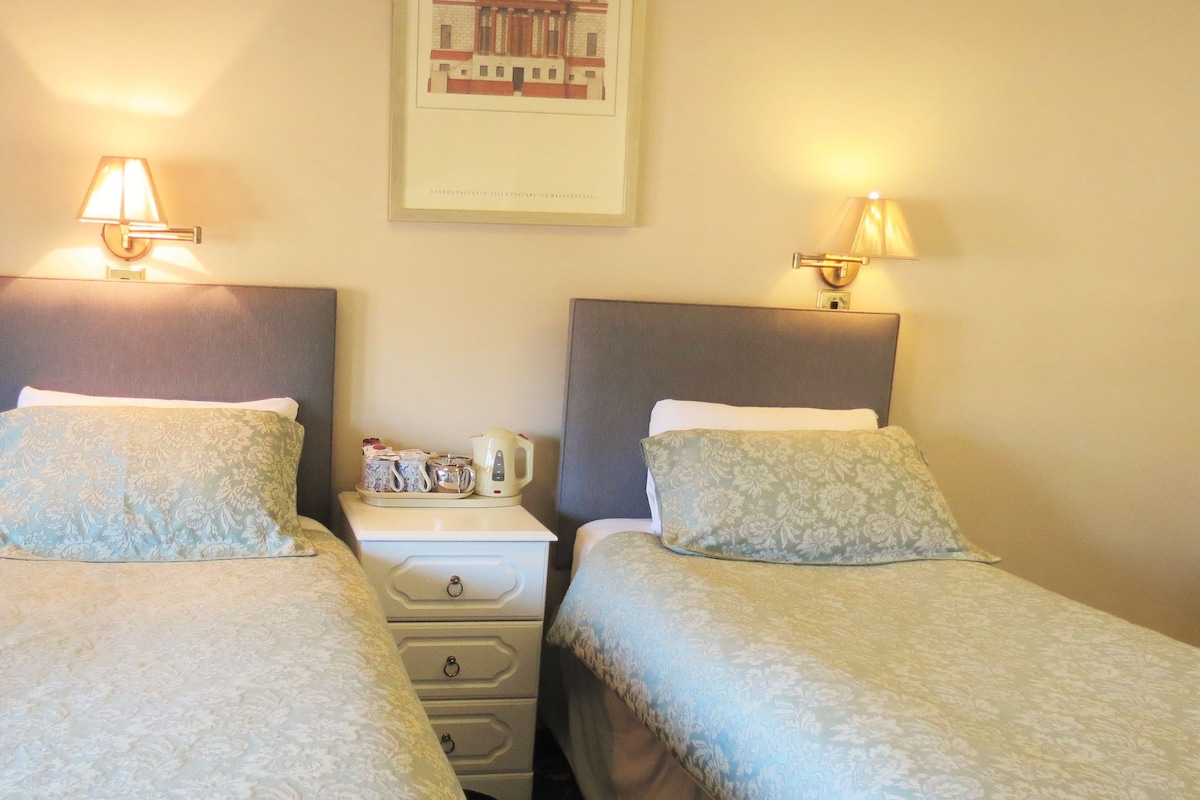 A hotel room with two beds, each with a headboard and bedside tables. There are lamps on the tables, a framed picture on the wall, and a clock on the wall.