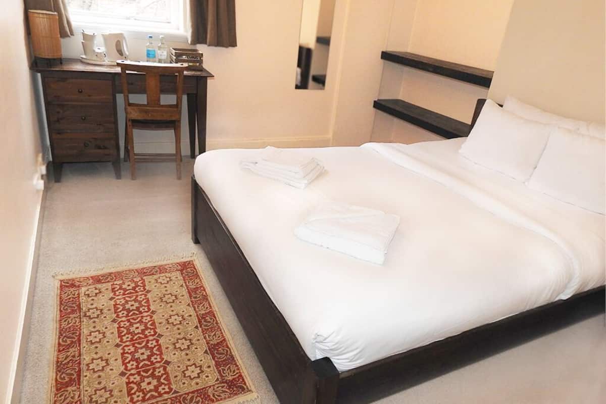 Una cama bien hecha con sábanas blancas, un escritorio de madera con una silla, una mesita con una lámpara y una alfombra estampada.