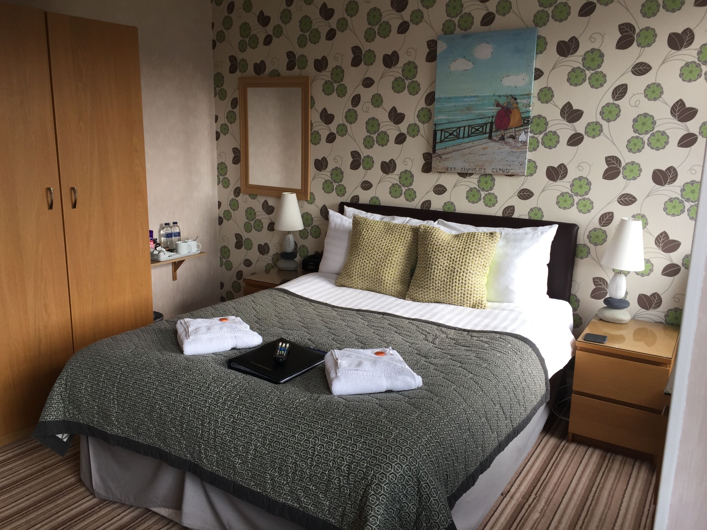 A hotel room with a bed, a nightstand, a wooden wardrobe, a painting, and a floral wallpaper.