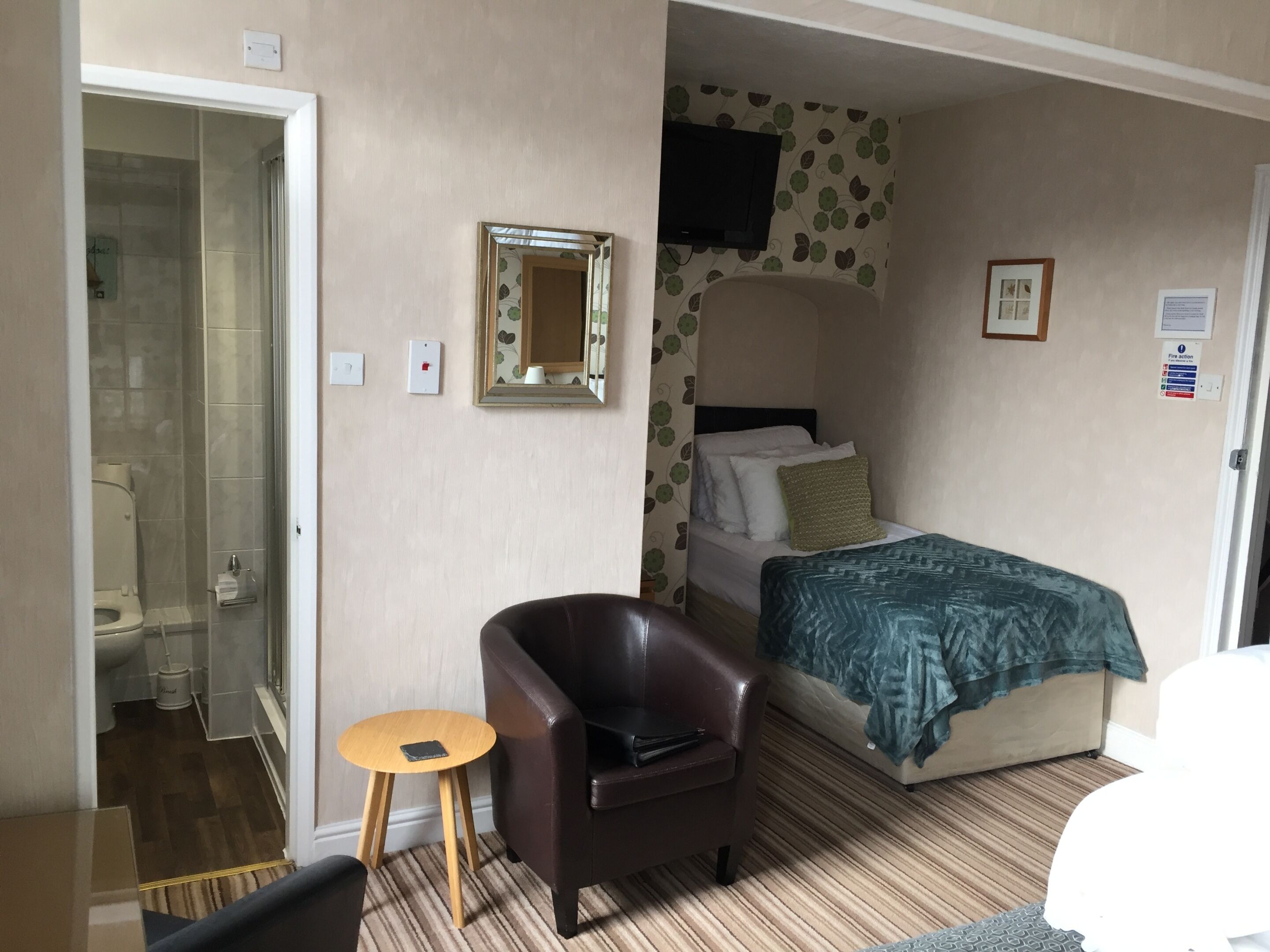 A hotel room with a bed, a chair, a small table, a mirror, and a shower area.