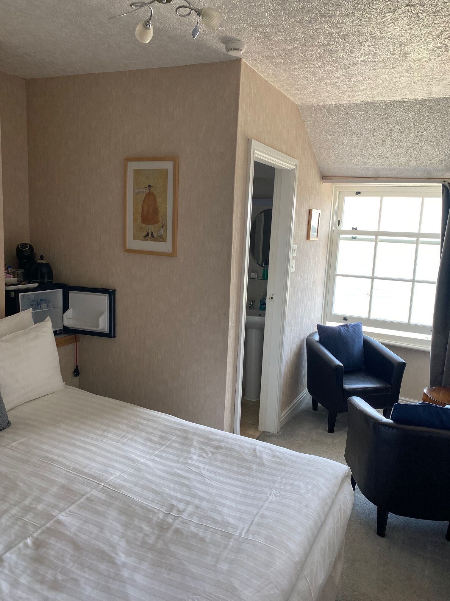 A hotel room with a large bed, two armchairs, a painting on the wall, and a window with a view.