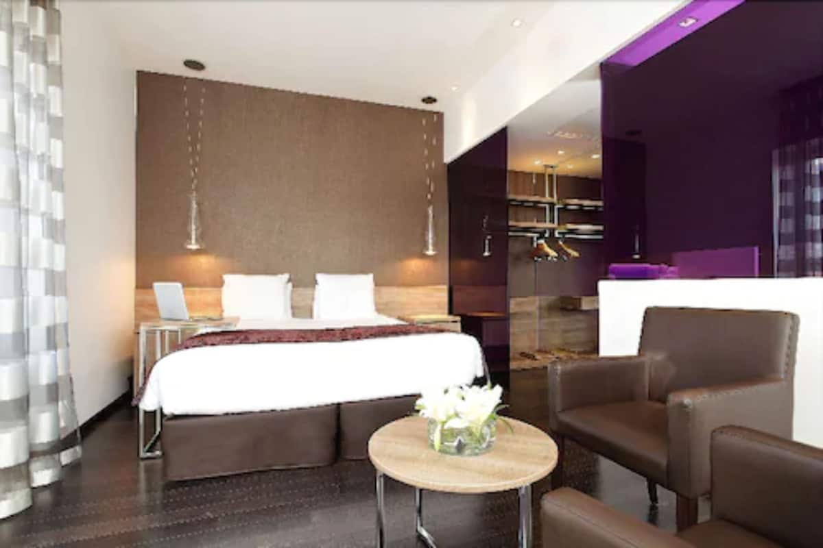 A modern hotel room with a bed, a small seating area, a round table with a vase, and a reception desk.