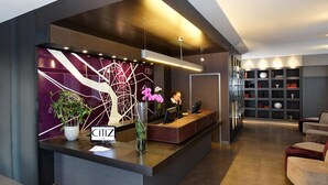 A modern hotel reception area with a dark reception desk, a large potted plant, and a decorative wall with a tree design. - Citiz Hotel (Toulouse)