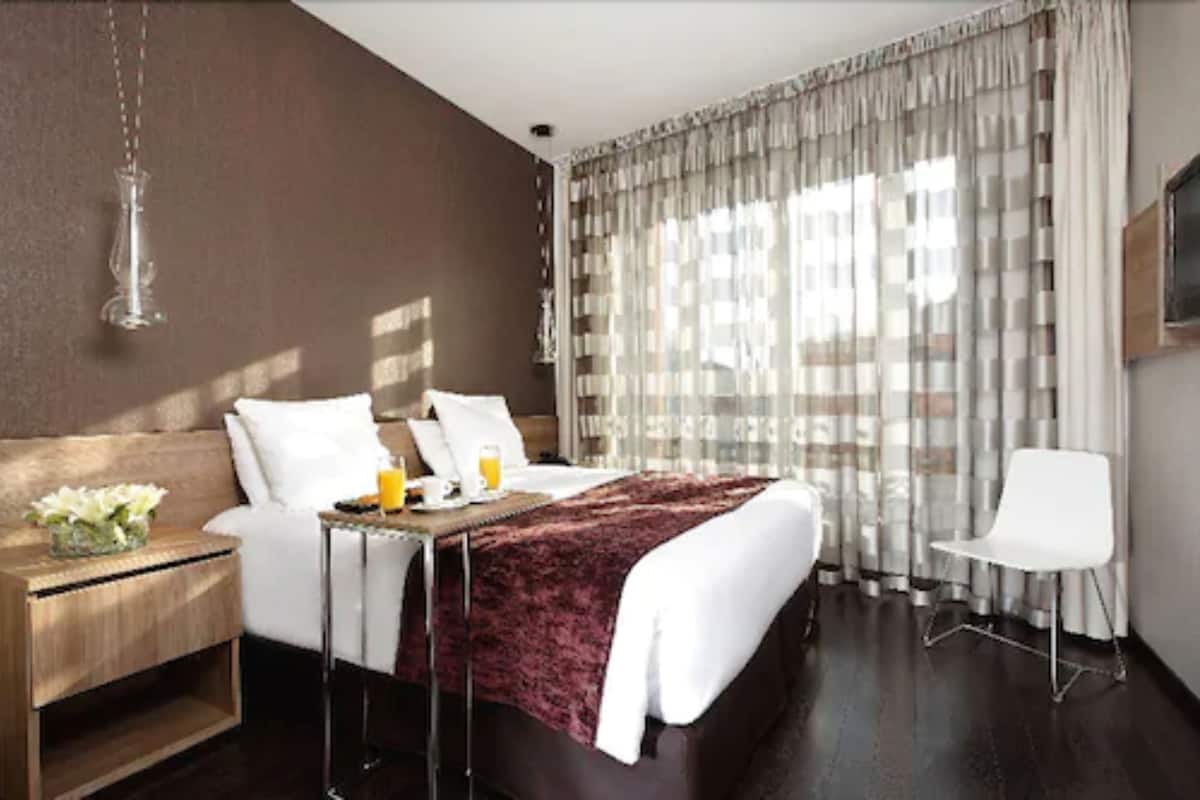 A modern hotel room with a large bed, a bedside table with a lamp, a chair, and a view of the outdoors through sheer curtains.