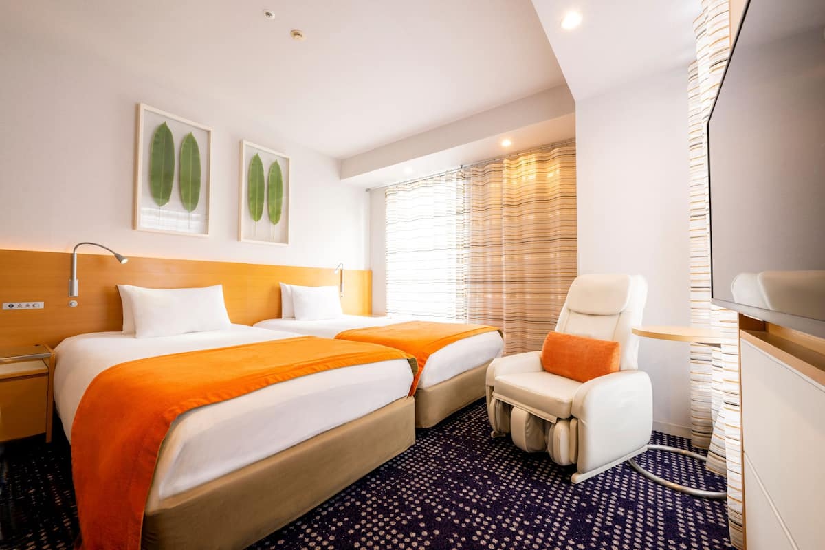 A hotel room with two beds, a massage chair, a TV, and a window with blinds.