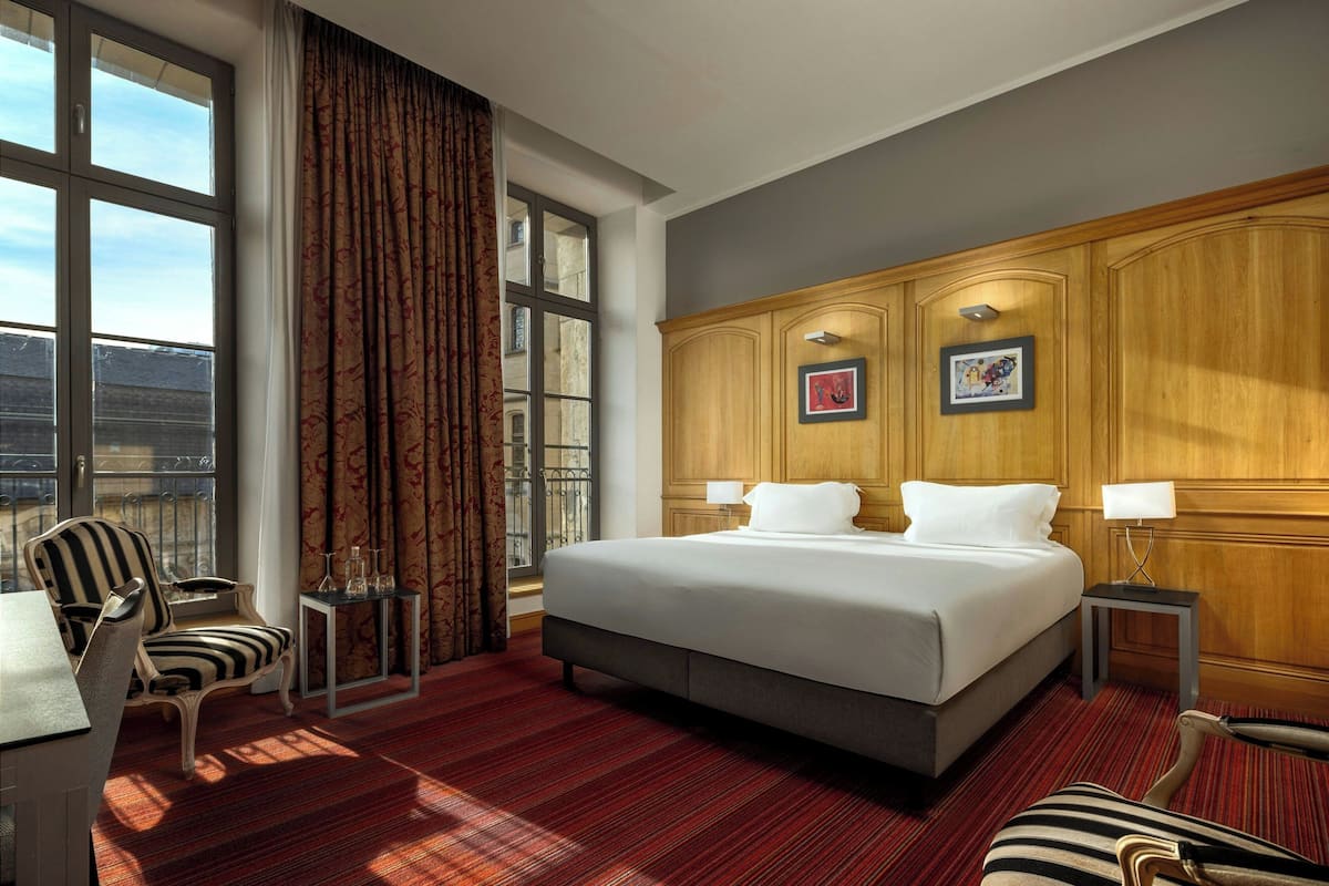 Habitación de hotel con una cama grande, dos mesitas de noche, una silla, una ventana con cortinas y vistas a edificios.