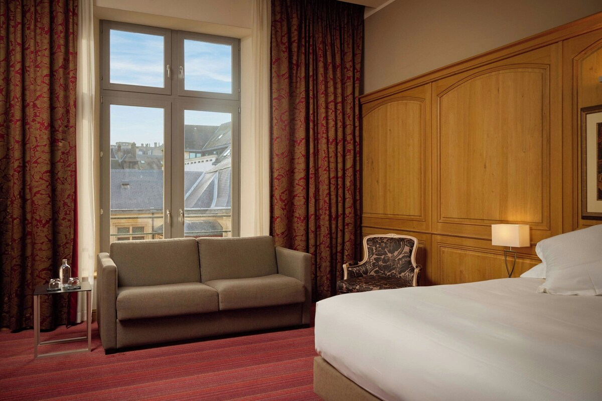 Une chambre d’hôtel avec un lit, un canapé, une chaise, une table et une fenêtre avec des rideaux.