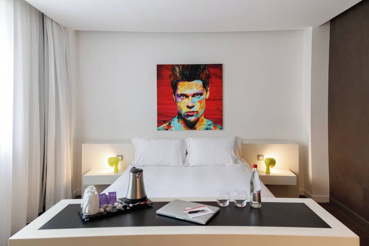 Habitación de hotel con cama, lámparas de noche, cafetera, laptop y un cuadro en la pared.