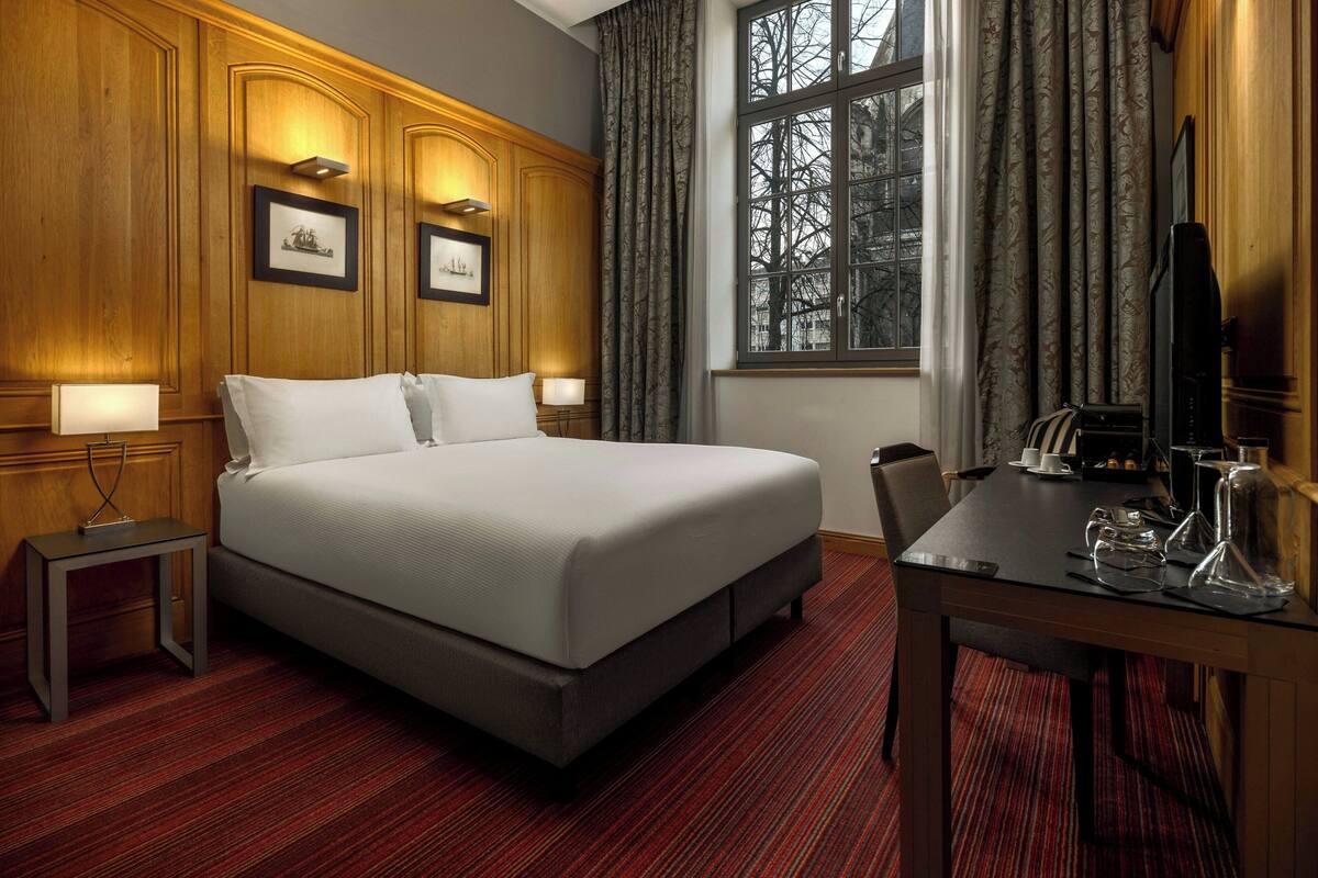 Habitación de hotel con una cama grande, una mesita con una silla, televisión y ventana con cortinas.