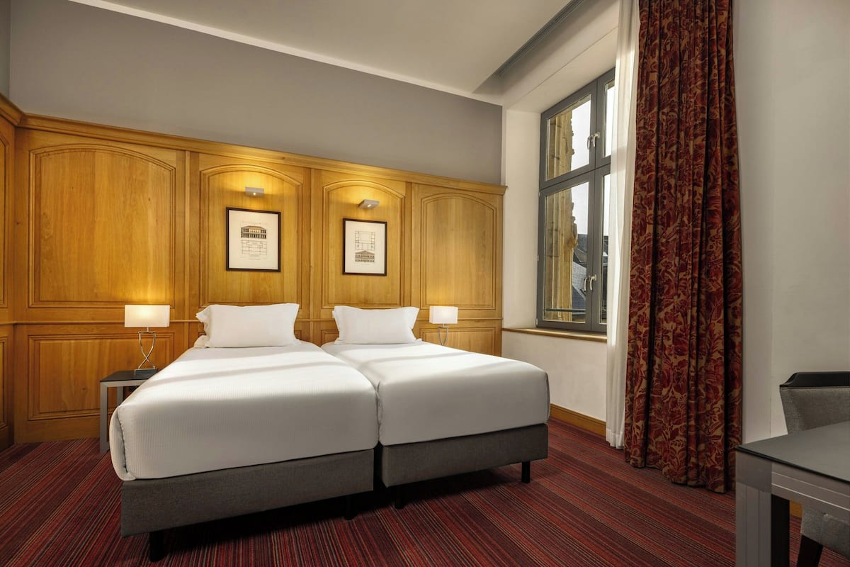 Habitación de hotel con dos camas, paneles de madera en las paredes, cuadros enmarcados, una ventana con cortinas y una pequeña mesa.