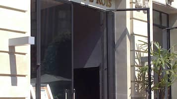 Uma área externa com assentos de vime, cadeiras e mesas, um tapete vermelho e a entrada de um edifício com detalhes ornamentados e uma placa com a inscrição "BRASSERIE DES 2 ROIS".