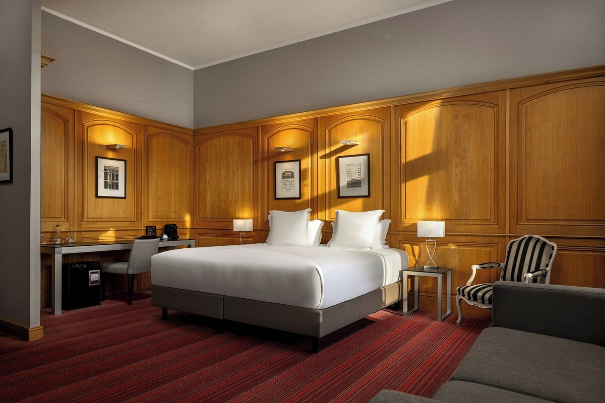Une chambre d’hôtel avec un grand lit, un bureau avec une chaise, une petite table avec une lampe et un fauteuil à rayures.