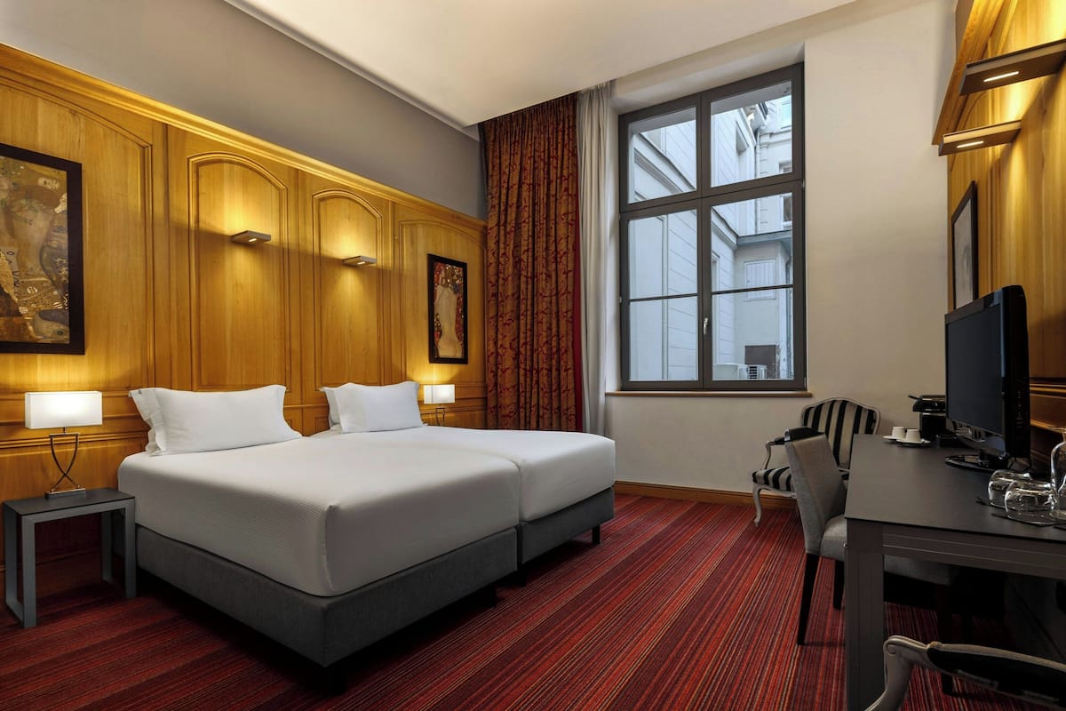Habitación de hotel con dos camas, un escritorio con computadora, una silla, un ventanal con vista a edificios y una alfombra roja.