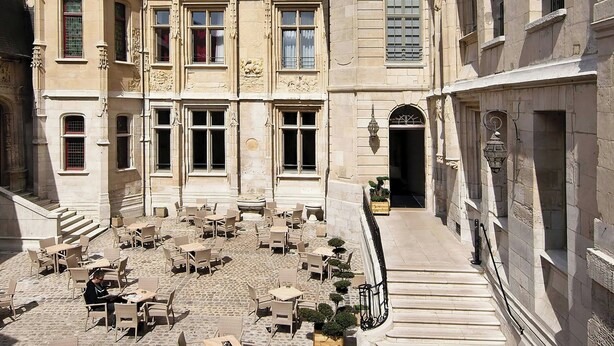 Une cour avec des tables et des chaises en pierre, un escalier menant à un niveau supérieur et un bâtiment doté d’une architecture gothique ornée.