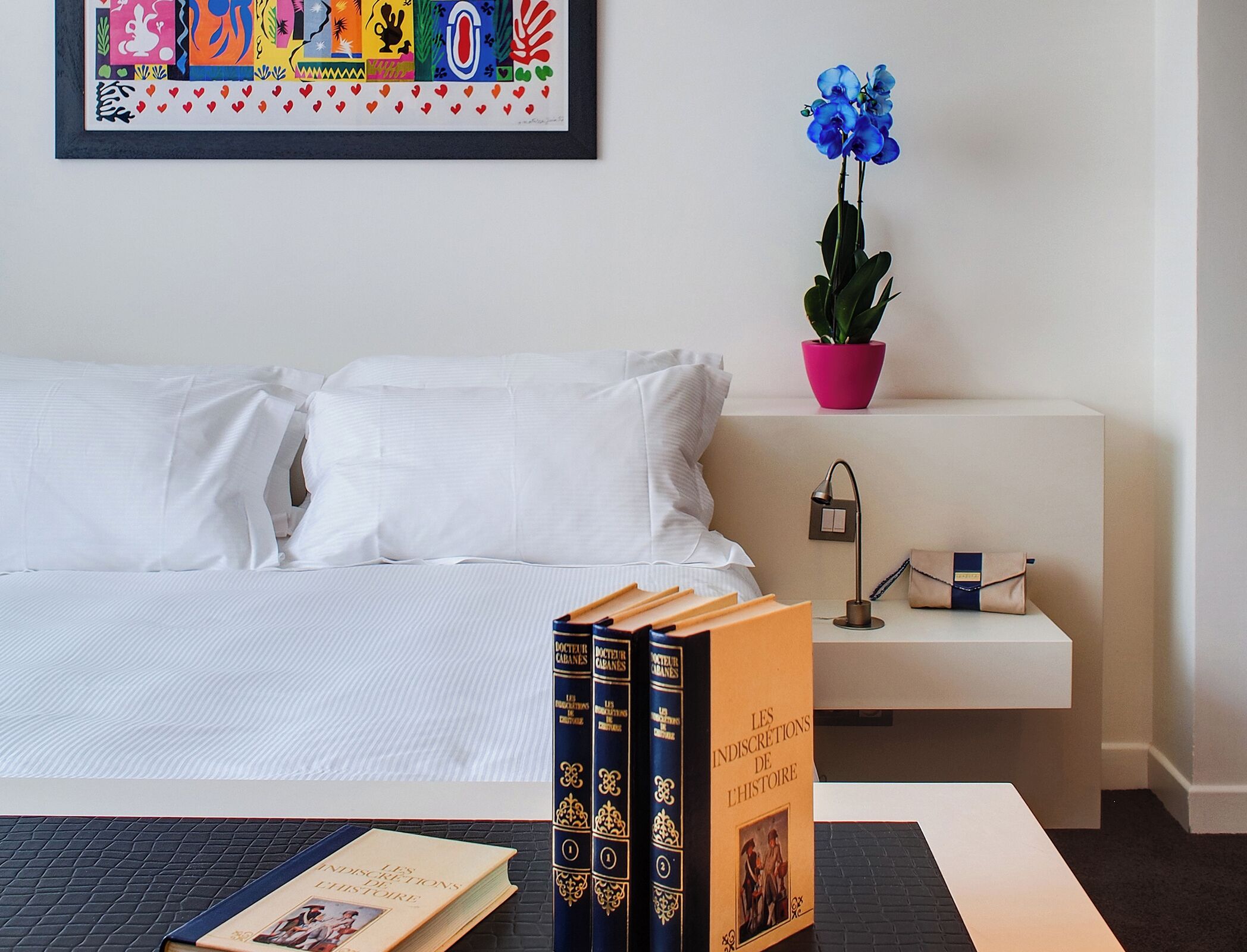 Een keurig opgemaakt slaapkamer met een bed, een nachtkastje met boeken, een lamp en een potplant.