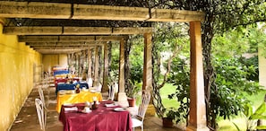 Un área de comedor al aire libre techada, con mesas dispuestas para una comida, rodeada de follaje y vistas despejadas al exterior.