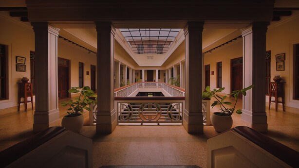 Un gran vestíbulo con una escalera central, flanqueado por columnas y plantas en macetas, que conduce a un techo alto con tragaluces.
