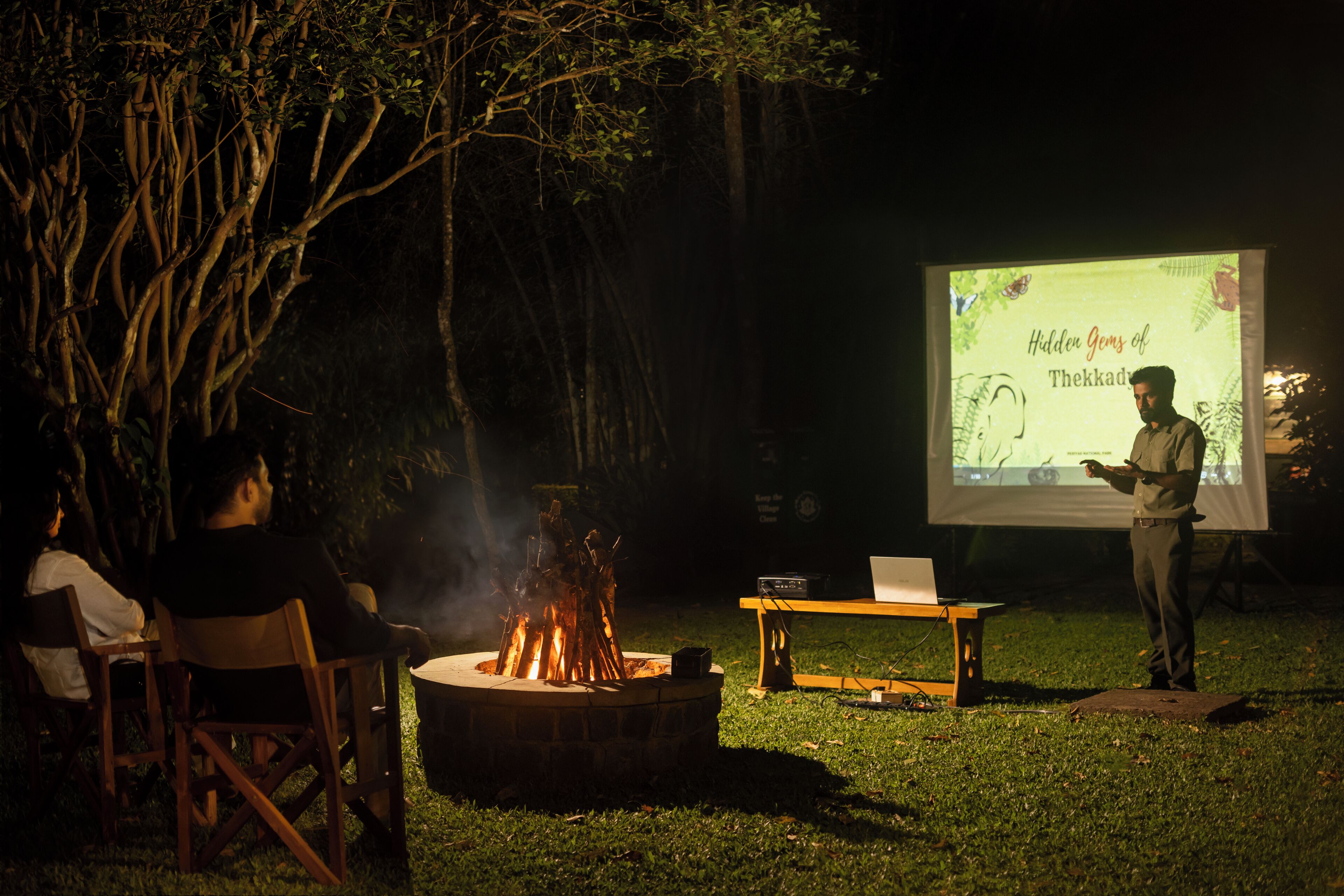 Presentación al aire libre con un brasero y una pantalla de proyector mostrando una presentación titulada 'Hidden Gems of Thekkad'.