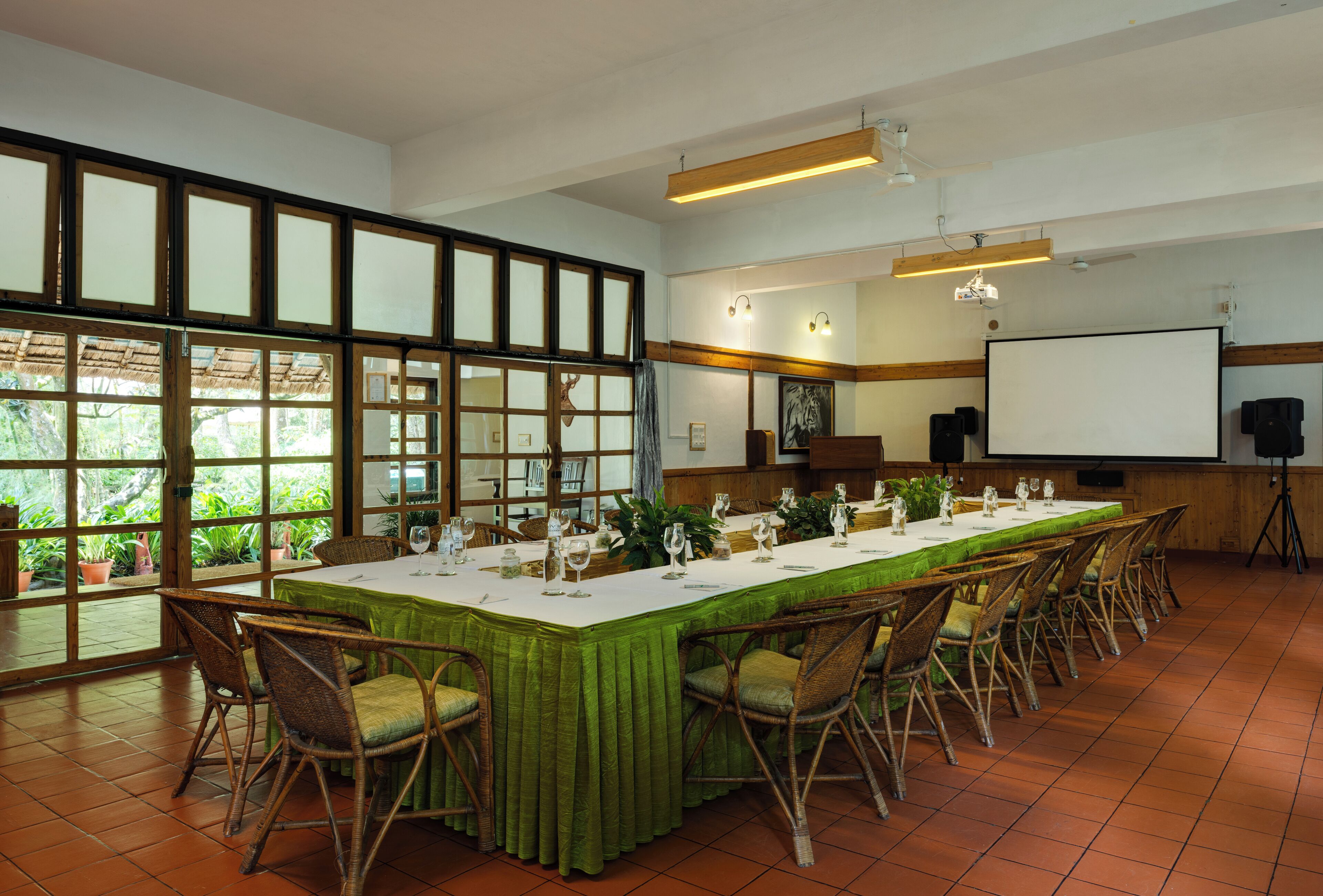 A conference room with a long table, chairs, a projector screen, and large windows with a view of greenery.