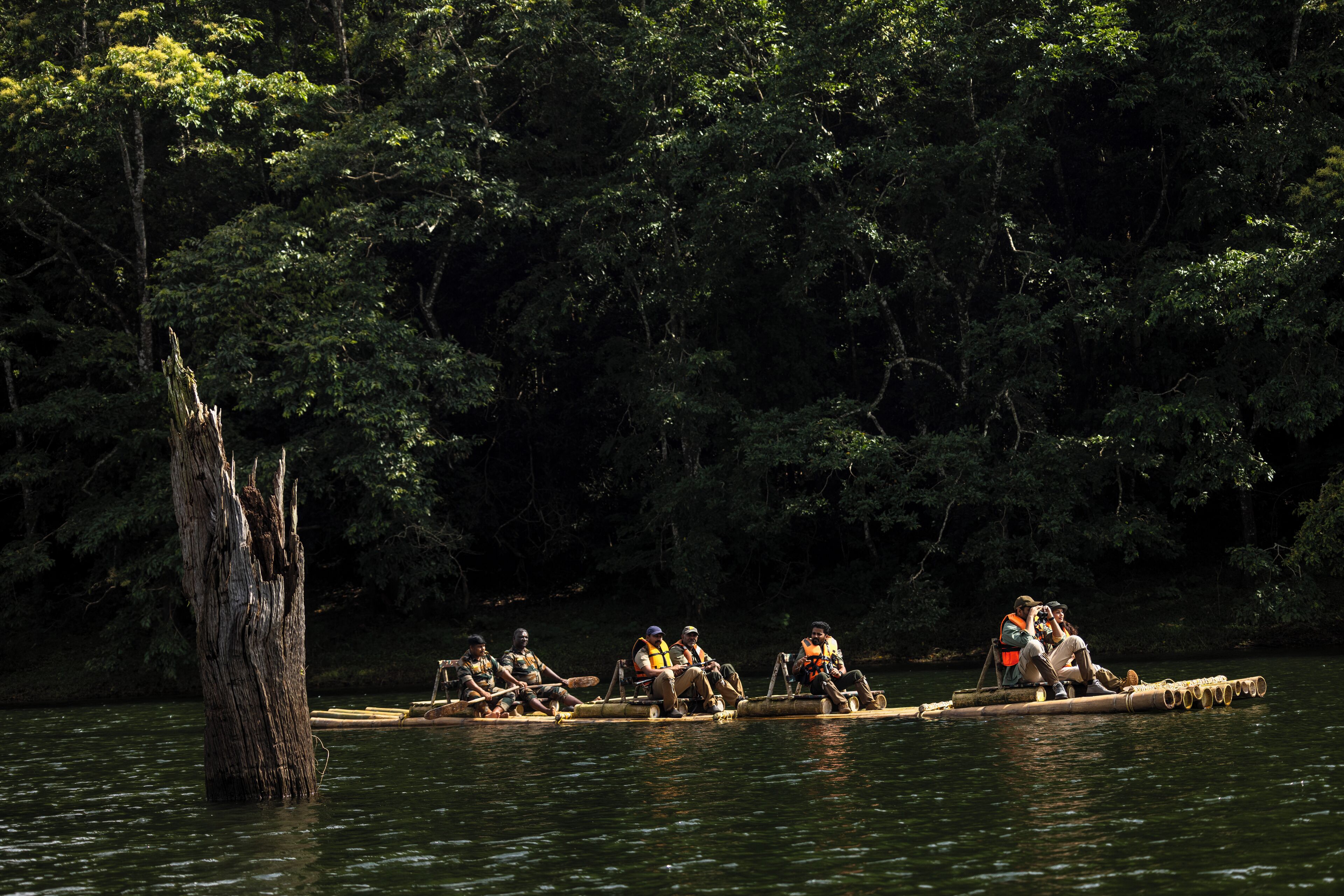 A group of people on a long, narrow boat on a river surrounded by dense forest.