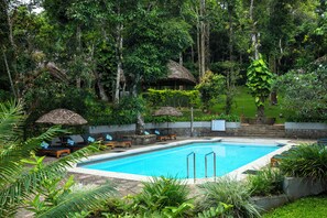 A swimming pool surrounded by lush greenery, with lounge chairs and thatched umbrellas. - Spice Village - Cgh Earth (Peermade)